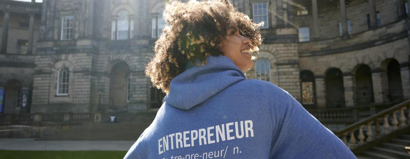 A young woman stands in front of Edinburgh's university building with her back to the camera, wearing a hoodie emblazoned with the word 'Entrepreneur'