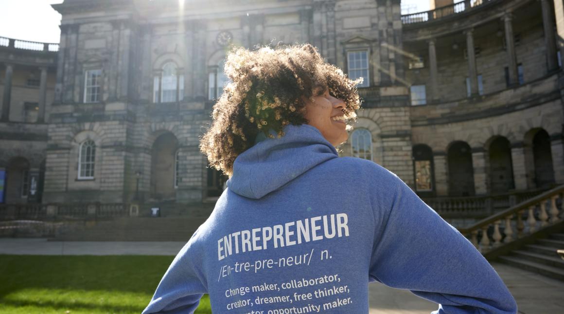 A young woman stands in front of Edinburgh's university building with her back to the camera, wearing a hoodie emblazoned with the word 'Entrepreneur'