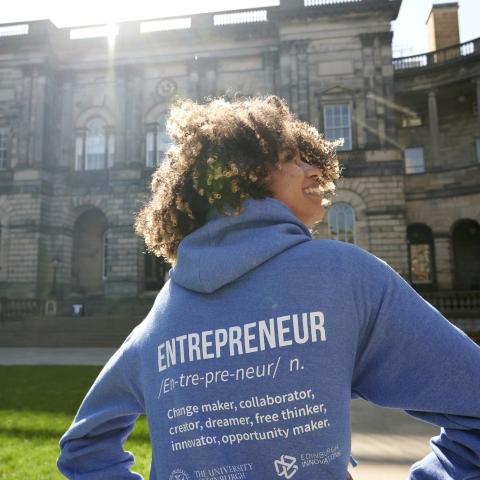 A young woman stands in front of Edinburgh's university building with her back to the camera, wearing a hoodie emblazoned with the word 'Entrepreneur'