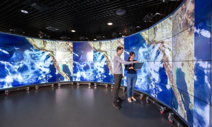 Researchers stand in front of large wall-to-wall digital screens displaying cloud patterns on global maps