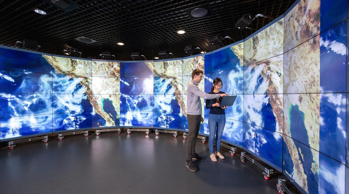Researchers stand in front of large wall-to-wall digital screens displaying cloud patterns on global maps