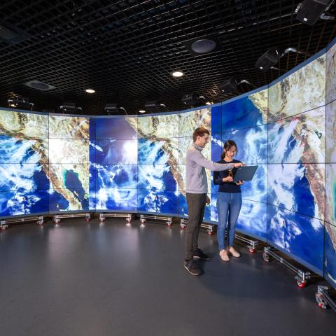 Researchers stand in front of large wall-to-wall digital screens displaying cloud patterns on global maps